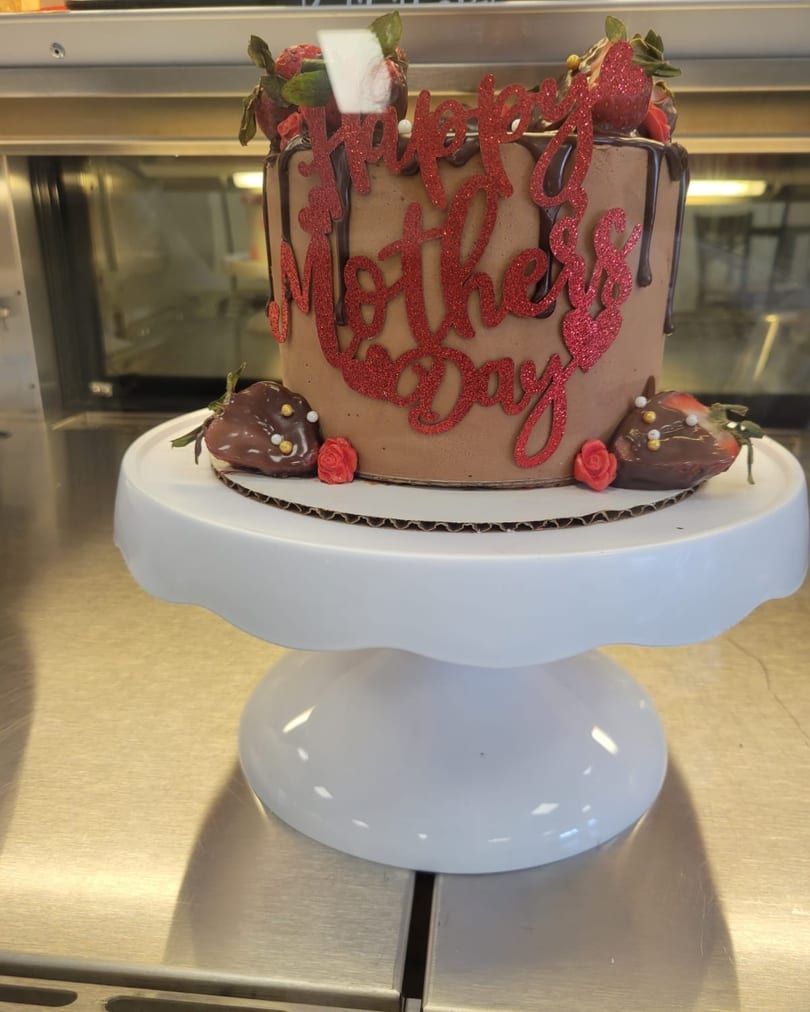 Chocolate Mother's Day cake with red glitter lettering, chocolate drizzle, and strawberries on a white stand.