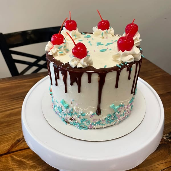 A Sweet Antojito cake with white frosting, chocolate drip, sprinkles, whipped cream, and cherries.