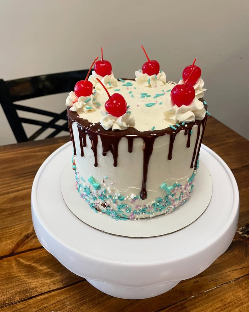 A Sweet Antojito cake with white frosting, chocolate drip, sprinkles, whipped cream, and cherries.