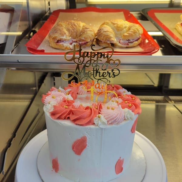 A white cake with pink frosting and a "Happy Mother's Day" topper, with croissants in the background.