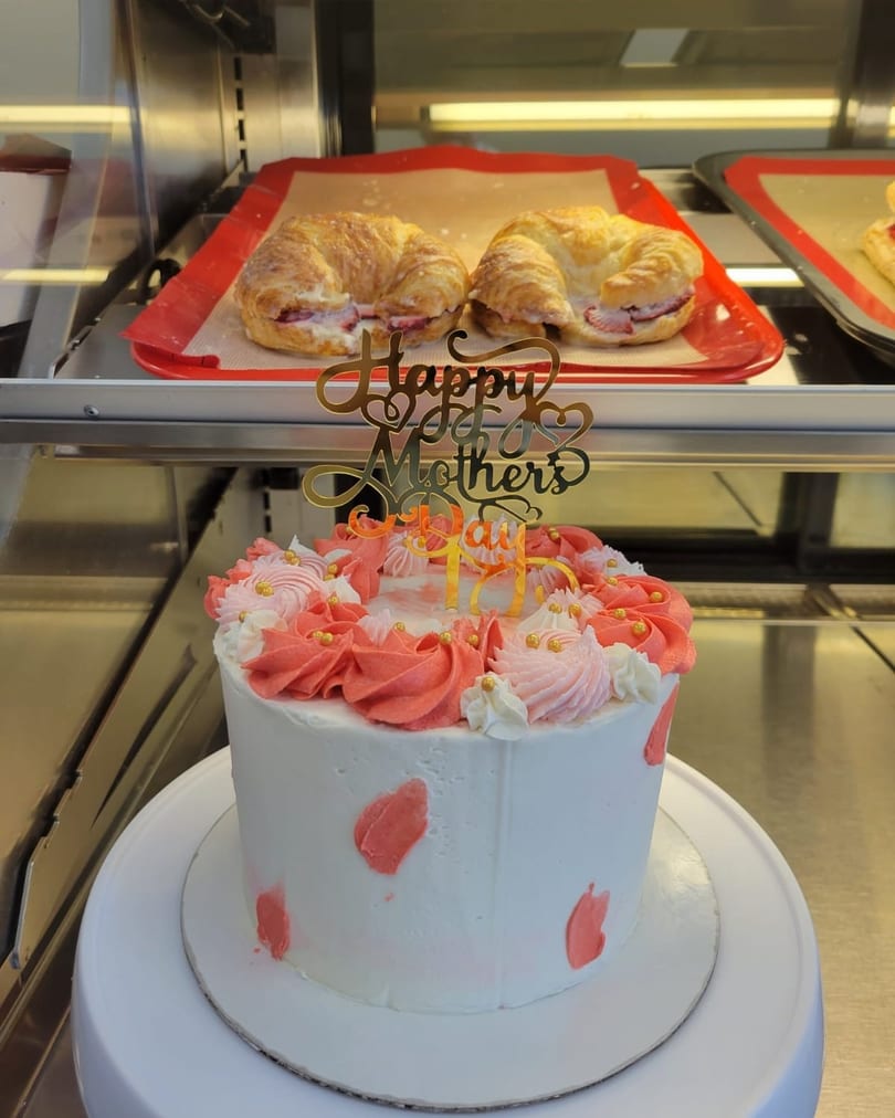 A white cake with pink frosting and a "Happy Mother's Day" topper, with croissants in the background.